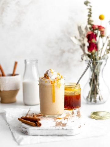 One vegan salted caramel white russian on a white cutting board in front of a jar of salted caramel, a jug of milk and a cup of cinnamon sticks.