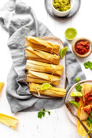 Platter of wrapped vegan sofritas tamales sat on a gray linen napkin next to small bowls of salsa and guacamole.