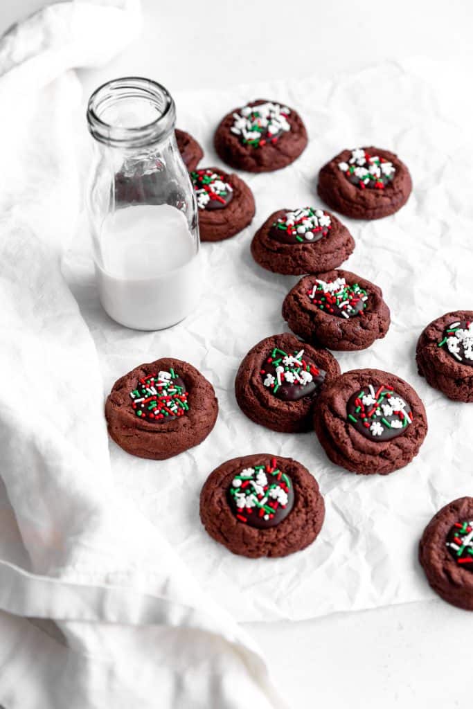 Vegan chocolate thumbprint cookies scattered across a white surface with a glass of milk and a linen napkin.