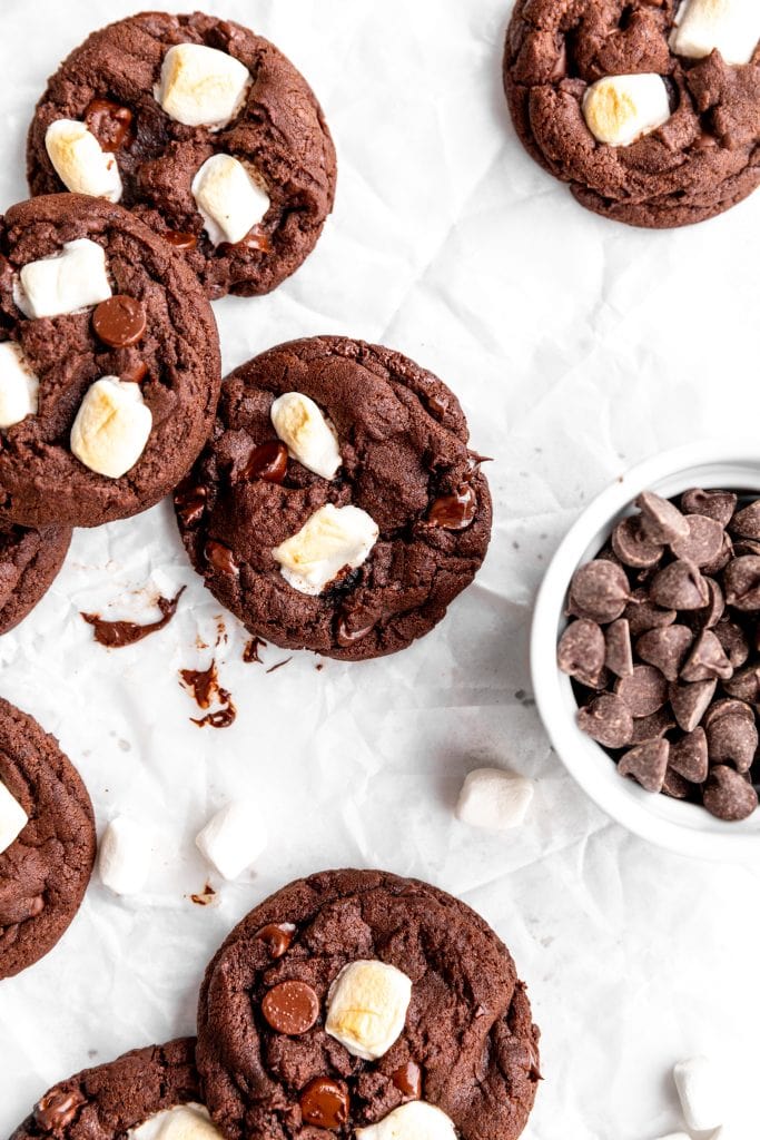 Vegan hot chocolate cookies spread across a white surface next to a cup of chocolate chips.