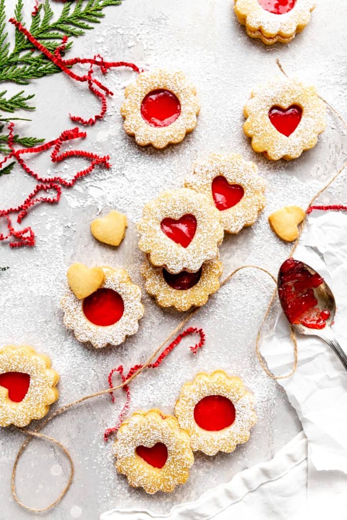 Vegan linzer cookies spread out on a gray surface next to a spoon covered in jam, a white linen napkin and a sprig of evergreen.