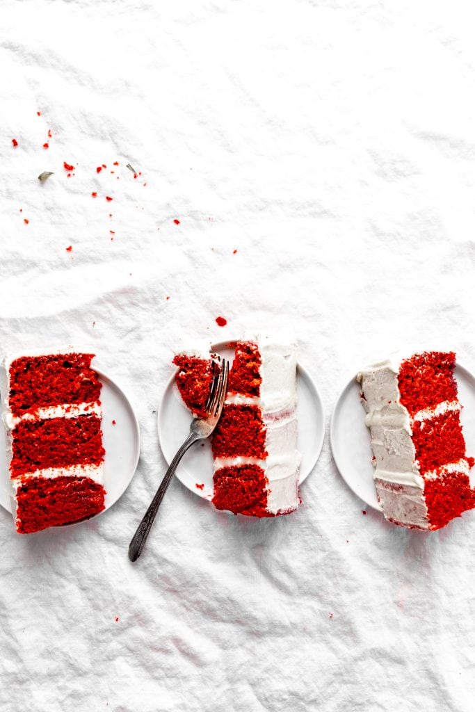 Three white plates with with slices of vegan red velvet cake sitting on a white linen tablecloth.