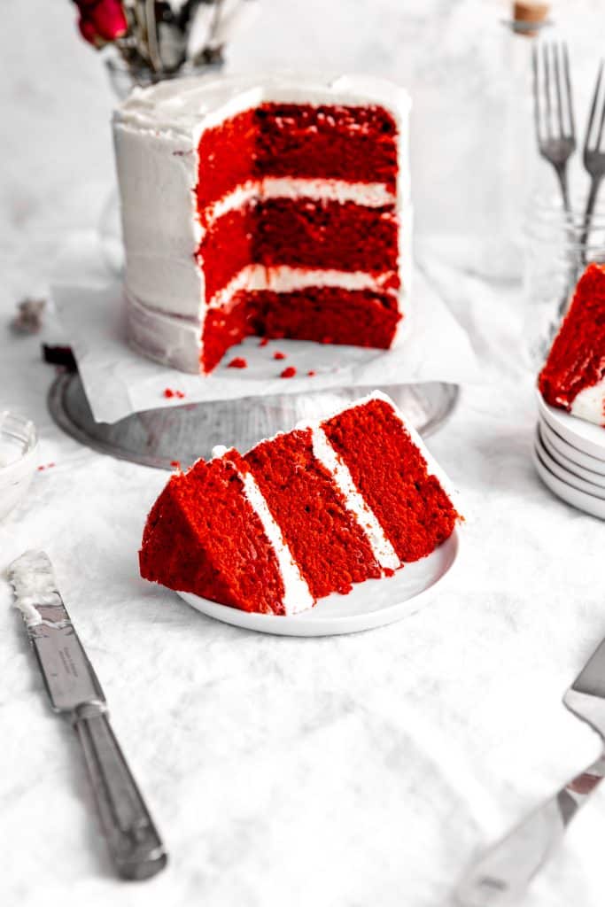 Slice of vegan red velvet cake on a white plate in front of the cake itself and a jar filled with forks.