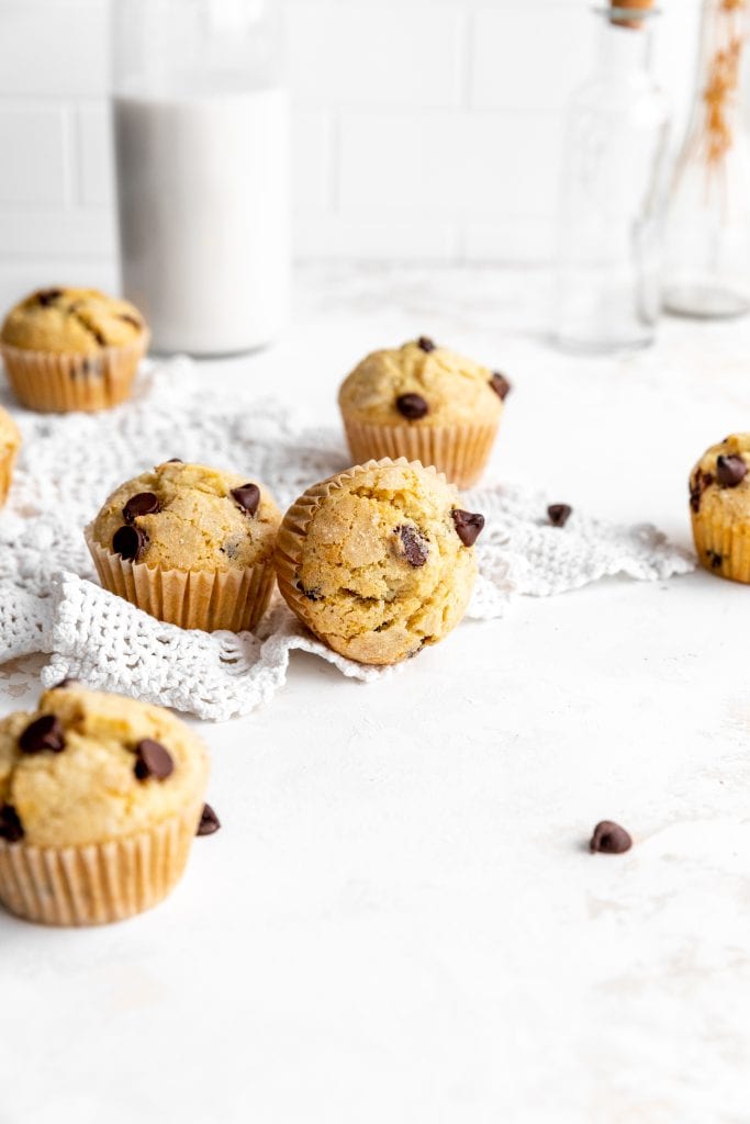 Vegan chocolate chip muffins scattered across a white surface in front of a jug of milk.