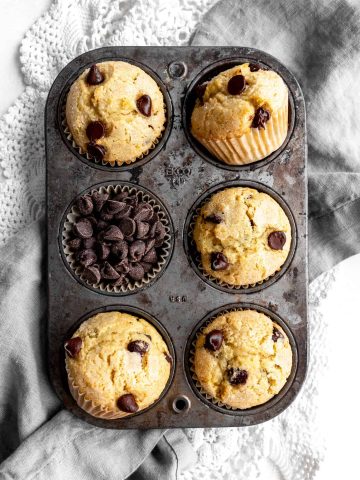 Baking pan filled with vegan chocolate chip muffins and loose chocolate chips sat on a gray napkin.