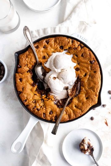 Two spoons digging into a skillet chocolate chip cookie topped with melty vanilla ice cream next to a glass of milk and stack of plates.