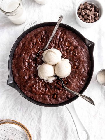 Multiple spoons digging into a vegan skillet brownie topped with vanilla ice cream.