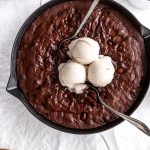 Multiple spoons digging into a vegan skillet brownie topped with vanilla ice cream.