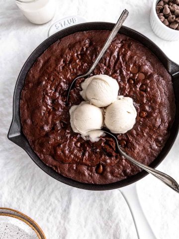 Multiple spoons digging into a vegan skillet brownie topped with vanilla ice cream.