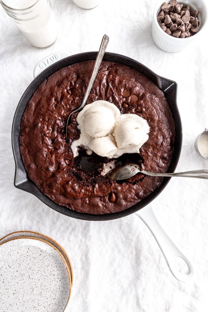 Multiple spoons digging into a vegan skillet brownie topped with melty vanilla ice cream.