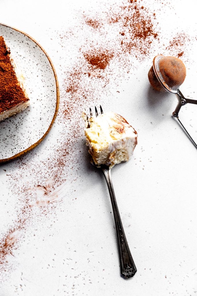 Fork holding a bite of tiramisu on a white background scattered with cocoa powder.