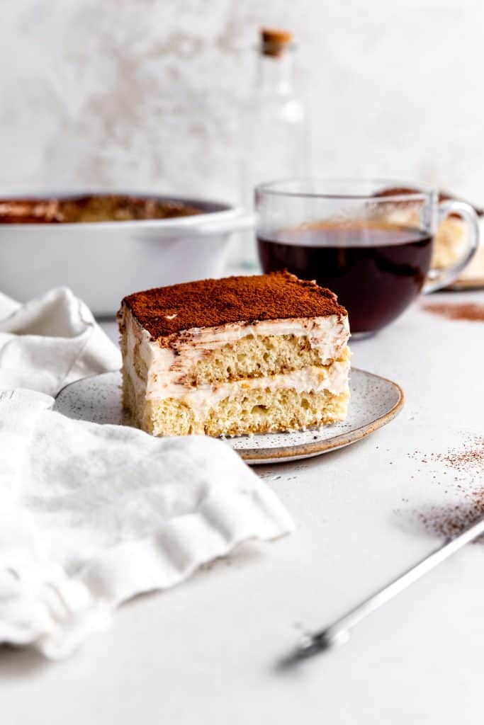 Slice of vegan tiramisu on a white plate in front of a cup of coffee, a white dish and a glass-corked bottle.