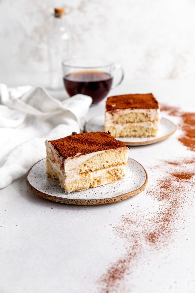 Two slices of vegan tiramisu on white plates in front of a cup of coffee.