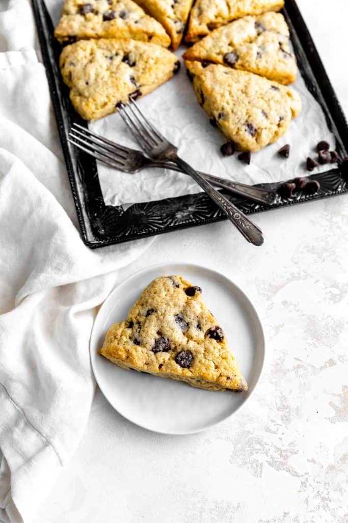 Chocolate chip scone on a white plate in front of baking sheet full of more scones.