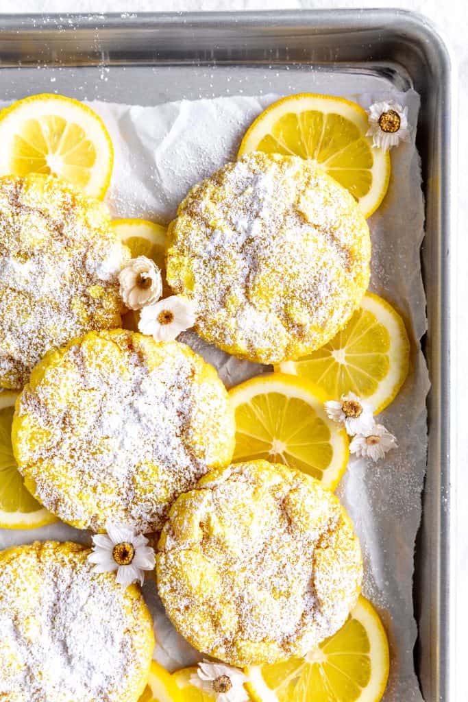 Lemon crinkle cookies on a baking sheet on white linen with lemon slices and flowers.