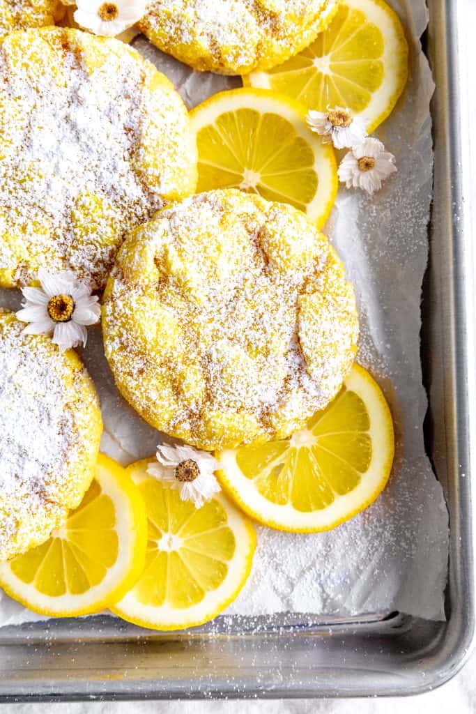 Lemon crinkle cookies on a baking sheet on white linen with lemon slices and flowers.