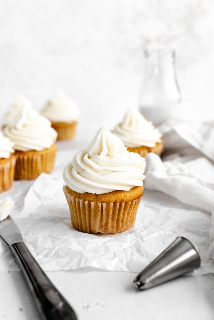 Vanilla cupcakes on a white surface with a jug of milk and a linen napkin.