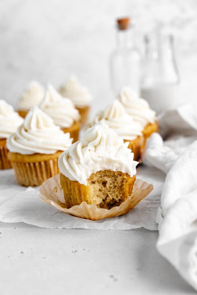 Vanilla cupcakes with a bite taken out of it in front of more cupcakes and a jug of milk.