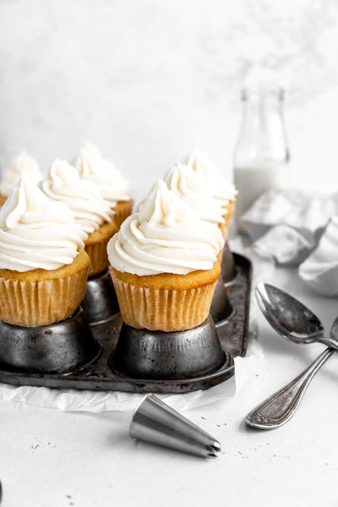 Vanilla cupcakes on a cupcake pan in front of a jug of milk.