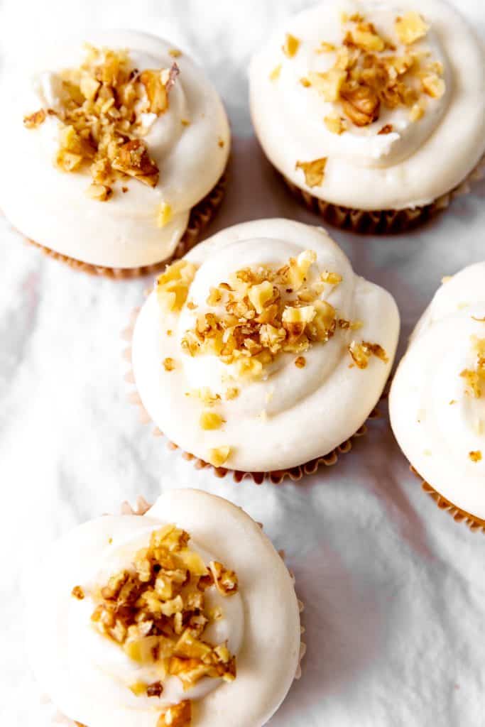 Tops of carrot cake cupcakes on a white linen surface with carrot shavings.