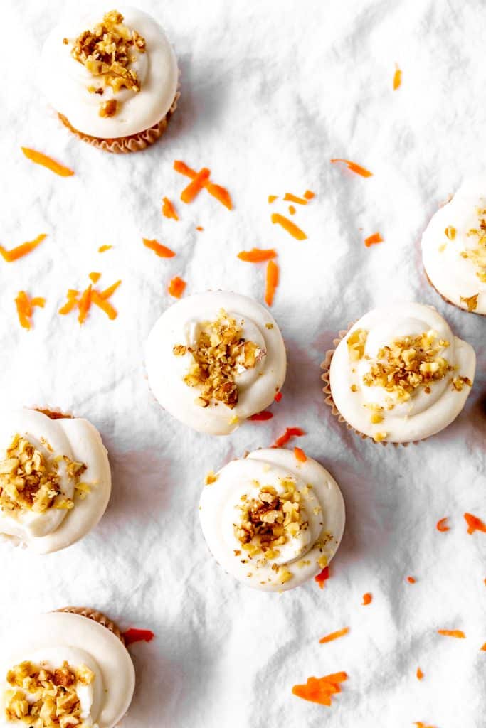 Tops of carrot cake cupcakes on a white linen surface with carrot shavings.