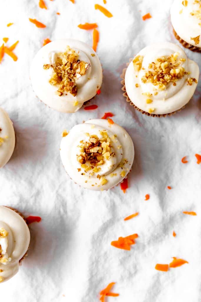 Tops of carrot cake cupcakes on a white linen surface with carrot shavings.