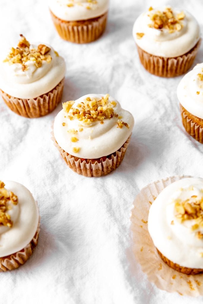 Carrot cake cupcakes on white linen topped with chopped walnuts.