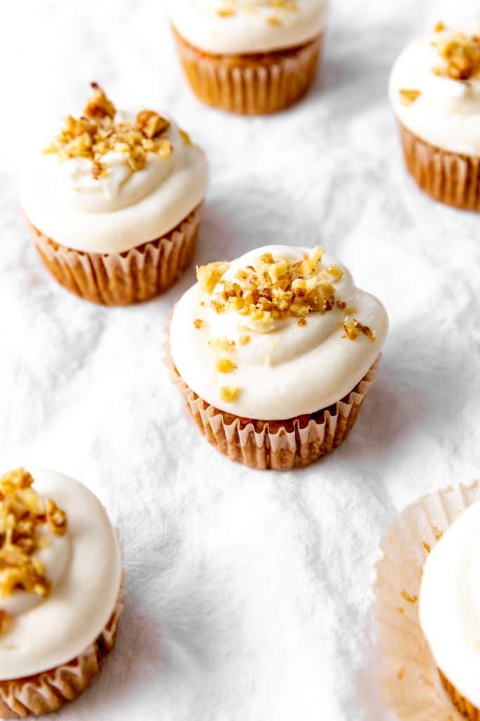 Carrot cake cupcakes on white linen topped with chopped walnuts.