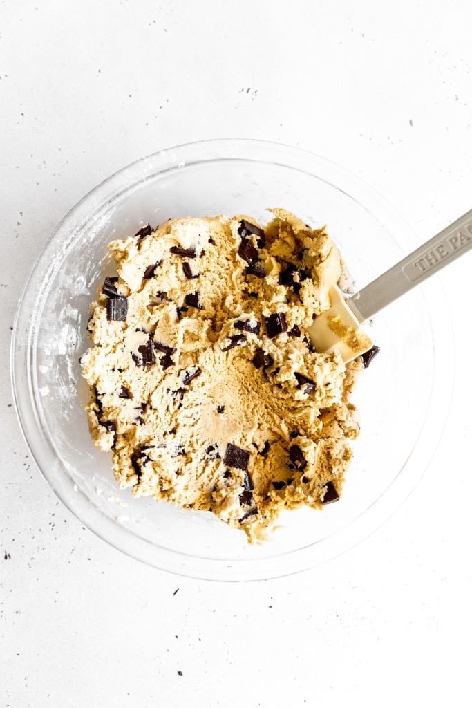 Chocolate chip cookie dough in a glass bowl with a rubber spatula.