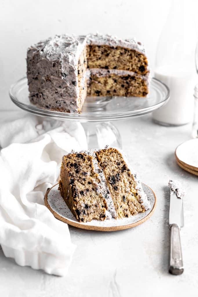 Slice or Oreo cake on a plate in front of the whole cake on a cake stand and a glass of milk.