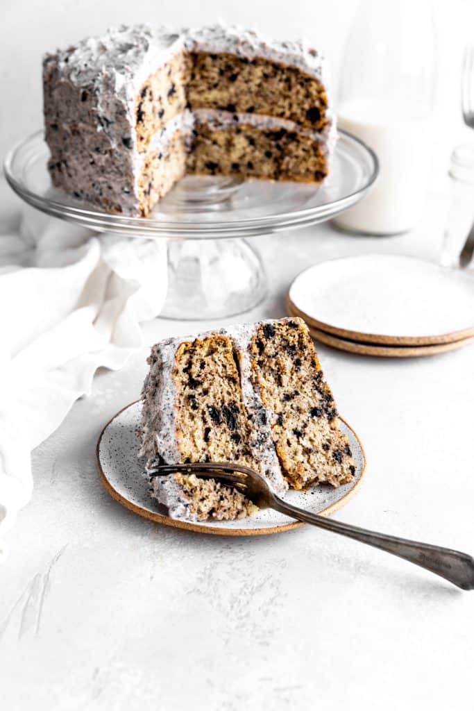 Fork cutting into a slice of Oreo cake on a white plate in front of the whole cake on a cake stand.