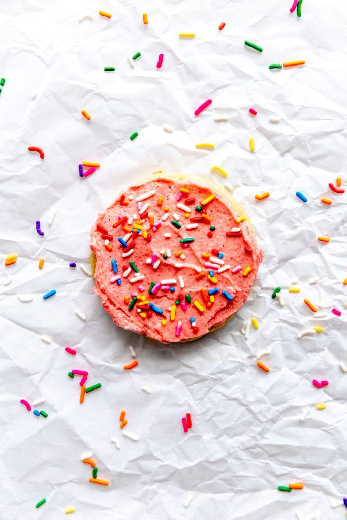 One pink frosted sugar cookie on a sheet of parchment paper covered in sprinkles.