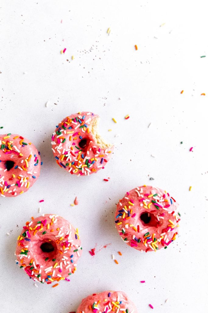 Baked donuts with pink icing and sprinkles on a white surface.