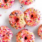 Baked donuts with pink frosting and sprinkles covering a white surface.