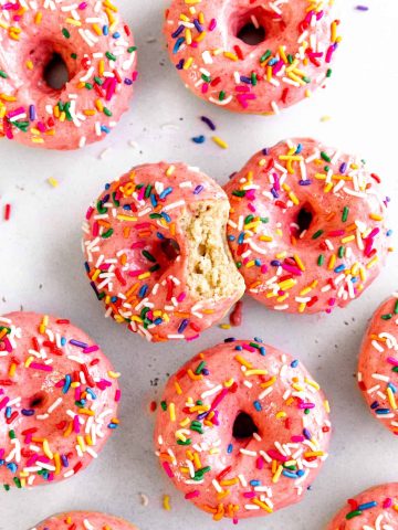 Baked donuts with pink frosting and sprinkles covering a white surface.