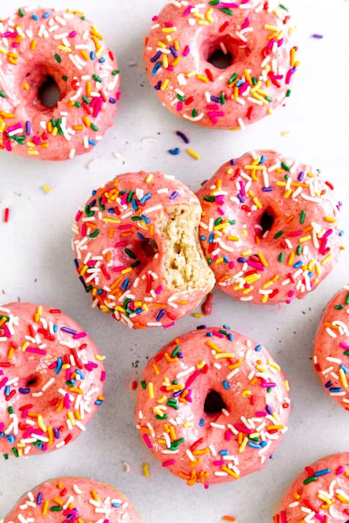 Baked donuts with pink frosting and sprinkles covering a white surface.