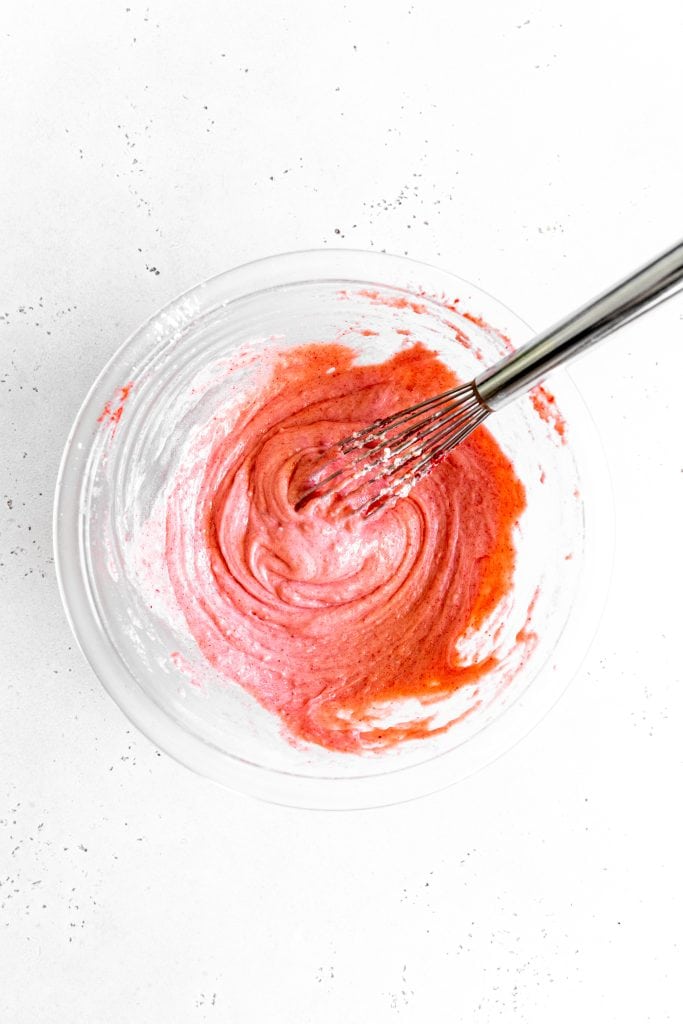 Glass bowl filled with pink strawberry icing and a whisk.