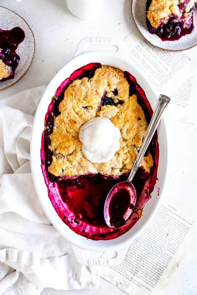 Spoon digging into a blackberry cobbler topped with vanilla ice cream.