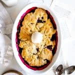 Blackberry cobbler in a baking dish topped with a scoop of vanilla ice cream next to a jug of milk.