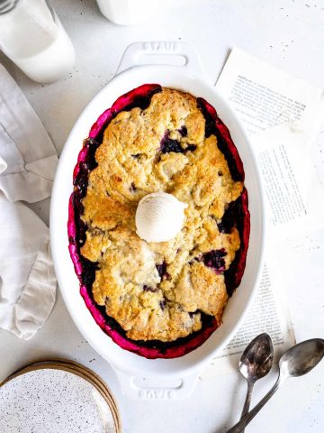 Blackberry cobbler in a baking dish topped with a scoop of vanilla ice cream next to a jug of milk.