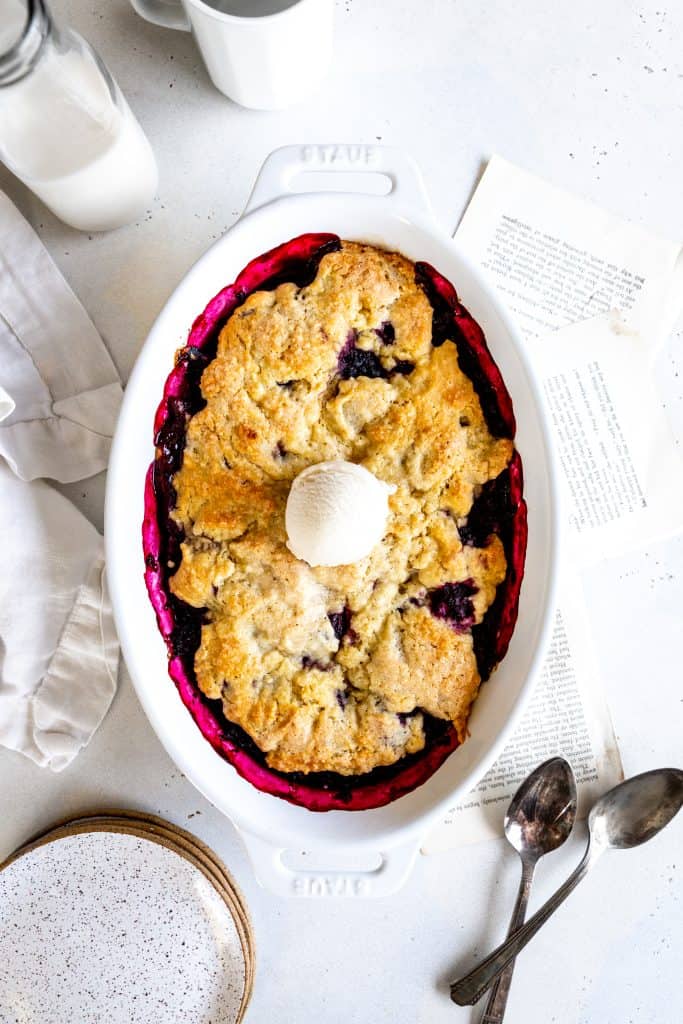 Blackberry cobbler in a baking dish topped with a scoop of vanilla ice cream next to a jug of milk.