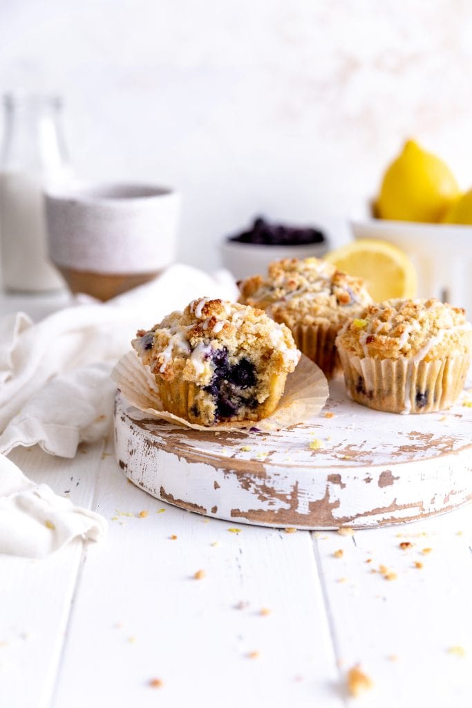 Lemon blueberry muffin with a bite taken out of it in front of more muffins, lemons and a jug of milk.