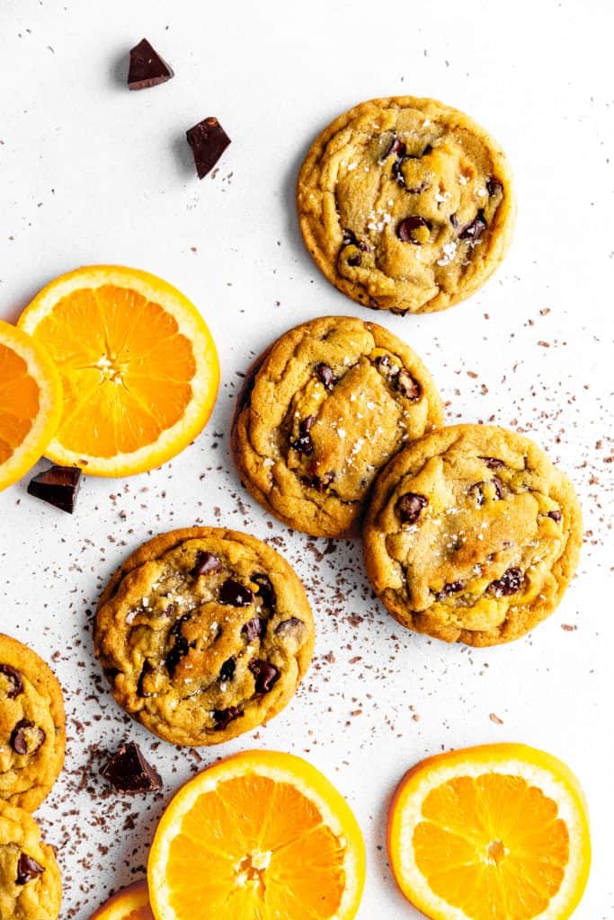 Orange chocolate chip cookies on a white surface with chocolate chunks and orange slices.