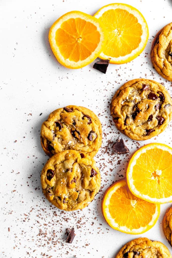 Orange chocolate chip cookies on a white surface with chocolate chunks and orange slices.