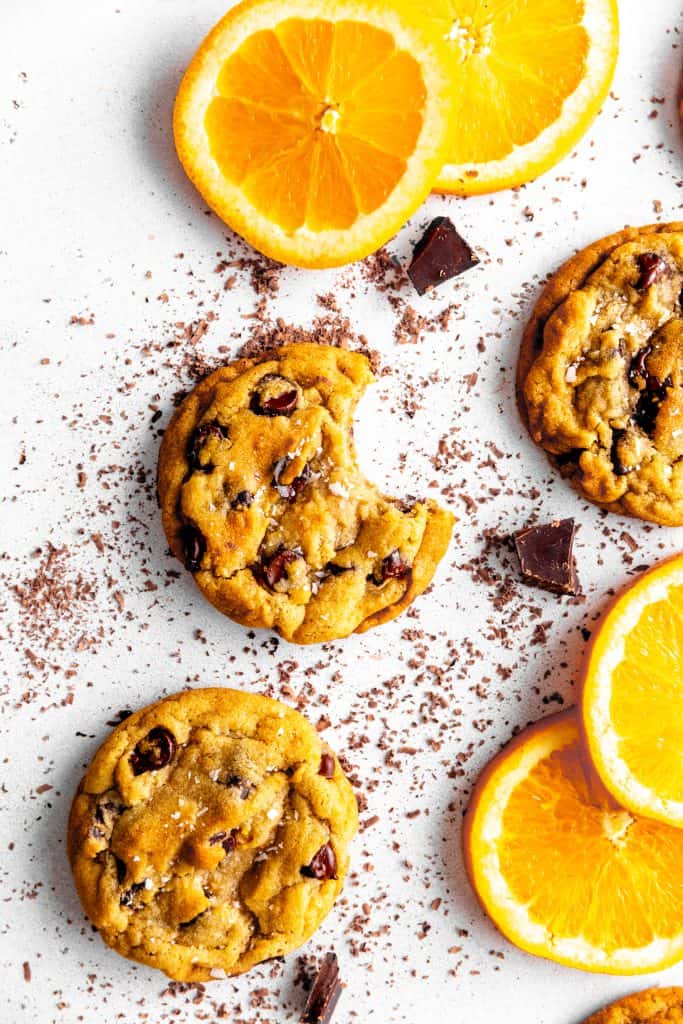 Orange chocolate chip cookie with a bite taken out of it surrounded by orange slices and chocolate chunks.