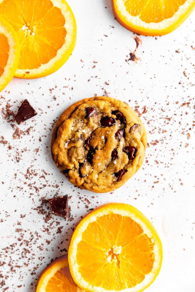 Orange chocolate chip cookie on a white surface covered in chocolate shavings and orange slices.