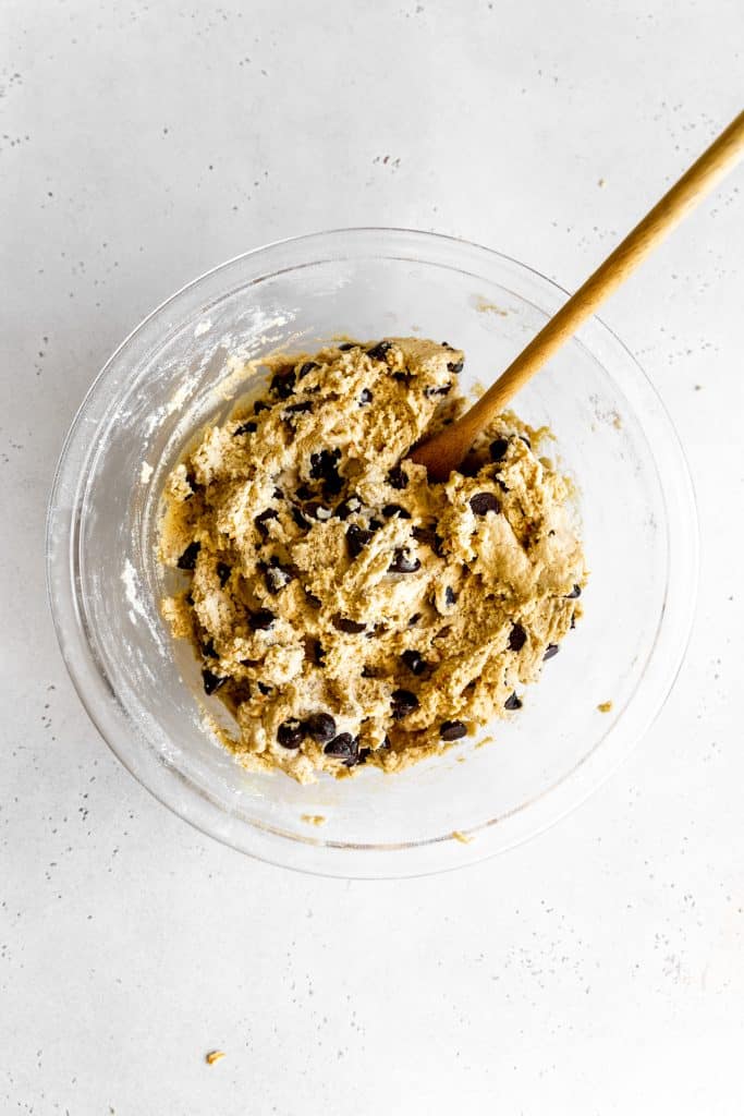 Glass bowl of chocolate chip cookie dough with a wooden spoon stuck in it.