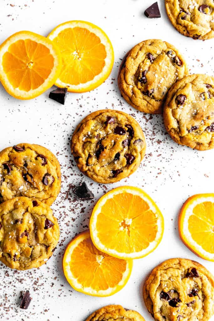 Orange chocolate chip cookies on a white surface with chocolate chunks and orange slices.