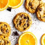 Orange chocolate chip cookies on a white surface with chocolate chunks and orange slices.