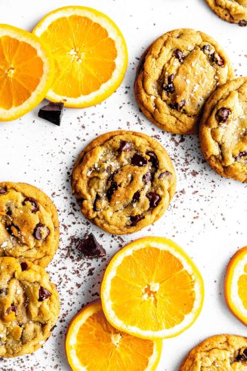 Orange chocolate chip cookies on a white surface with chocolate chunks and orange slices.